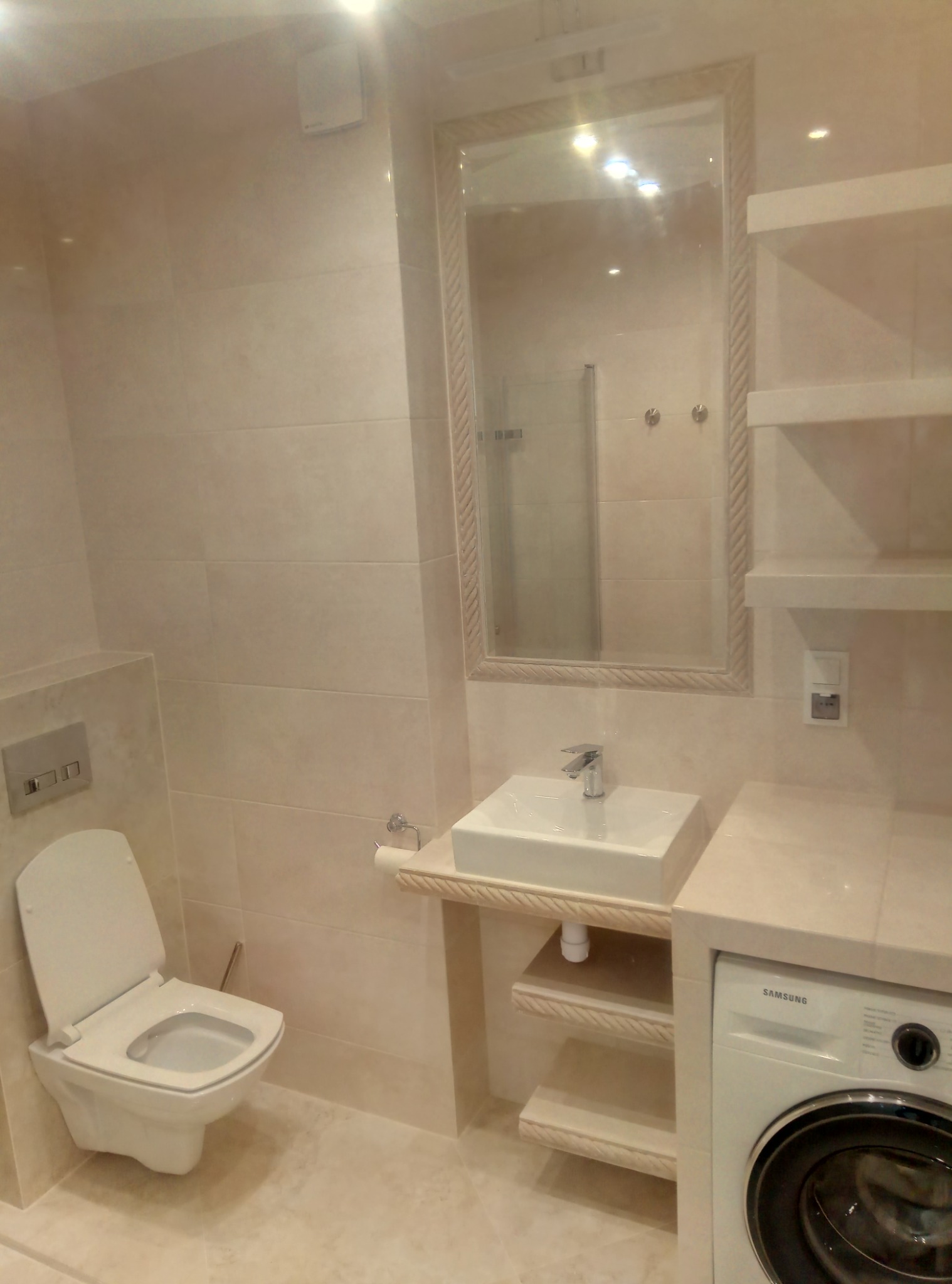 Jasna łazienka po remoncie: podwieszana toaleta, umywalka z szafką i lustrem w ozdobnej ramie oraz pralka Samsung w zabudowie.