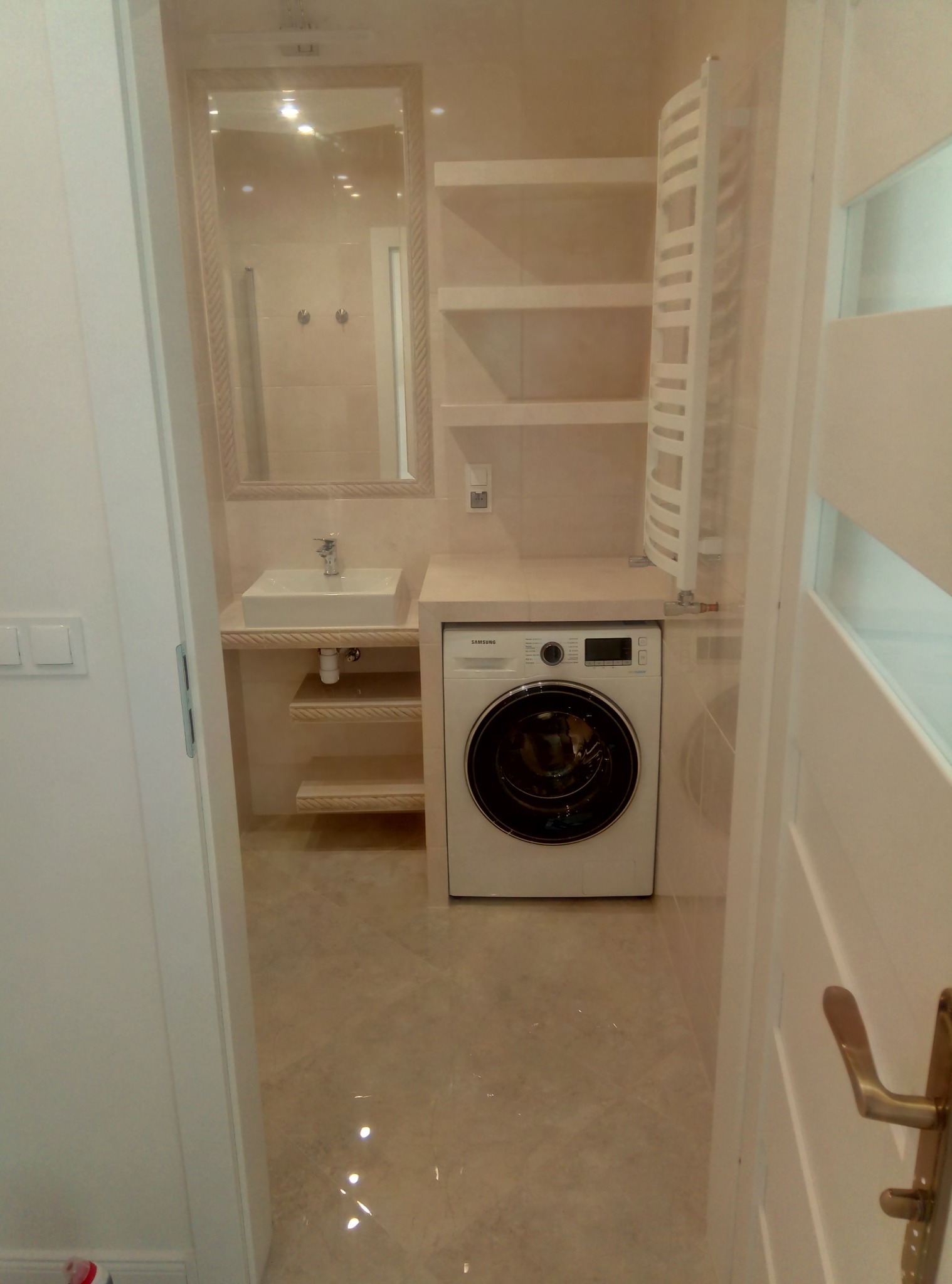 Beżowa łazienka z pralką zabudowaną pod blatem z umywalką, lustro w ramie, grzejnik drabinkowy i półki, widok zza uchylonych drzwi.