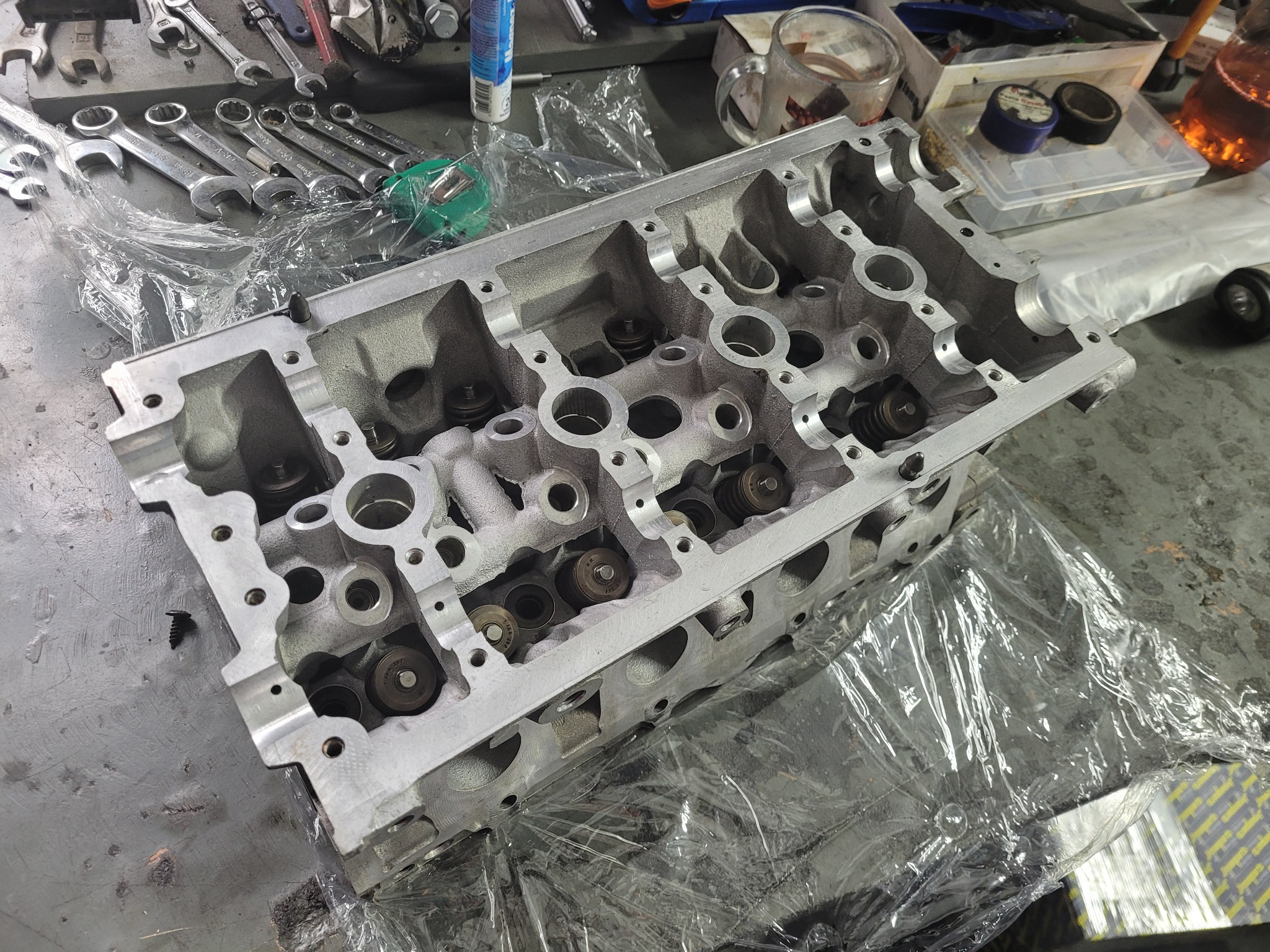 Głowica silnika 2.0 TFSI na warsztatowym stole, widoczne zawory, sprężyny, narzędzia i opakowanie folii ochronnej.