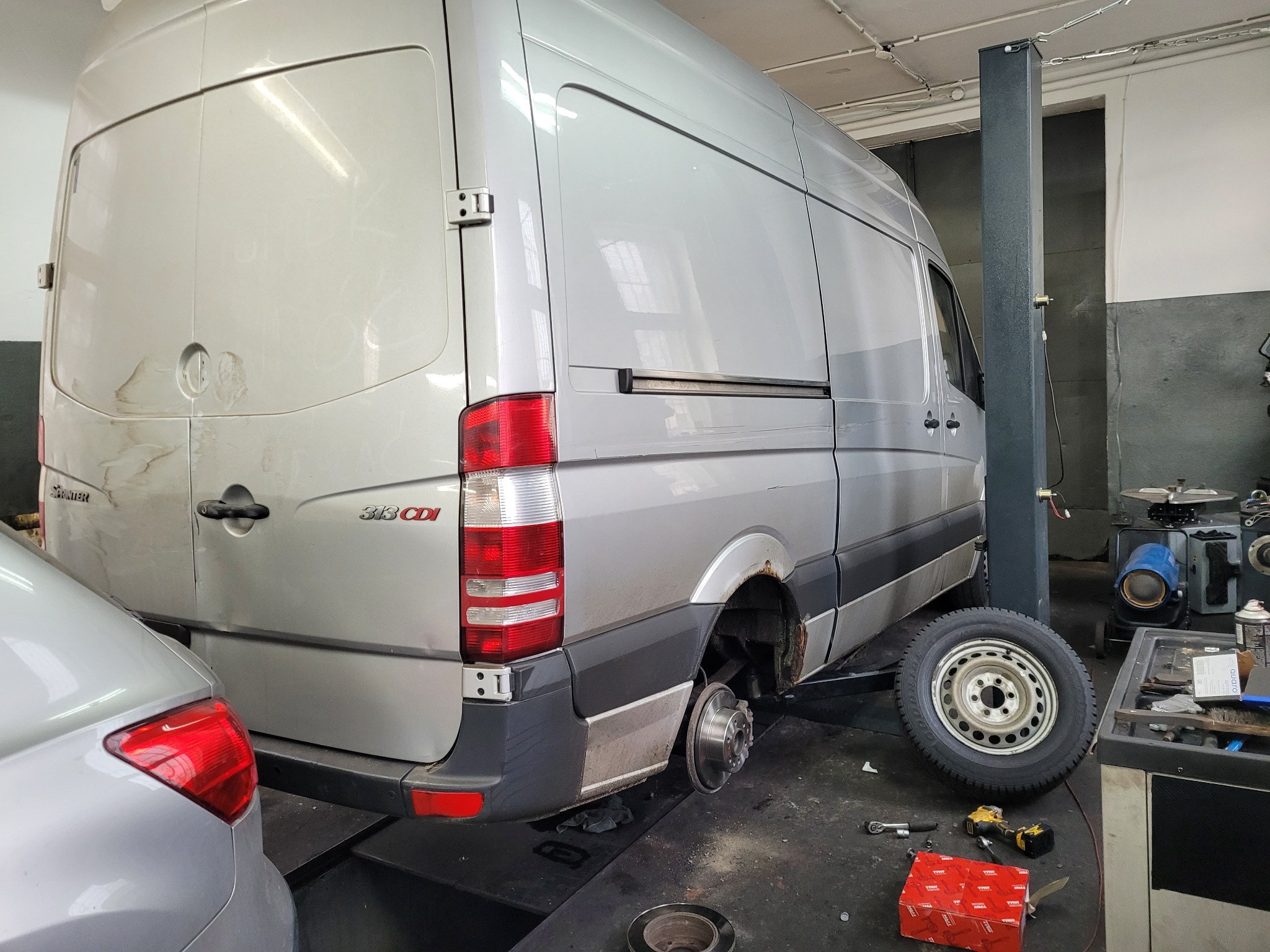Srebrny bus Mercedes Sprinter 313 CDI na podnośniku w warsztacie, zdemontowane tylne koło, widoczna korozja nadkoli, obok leży opona i narzędzia.