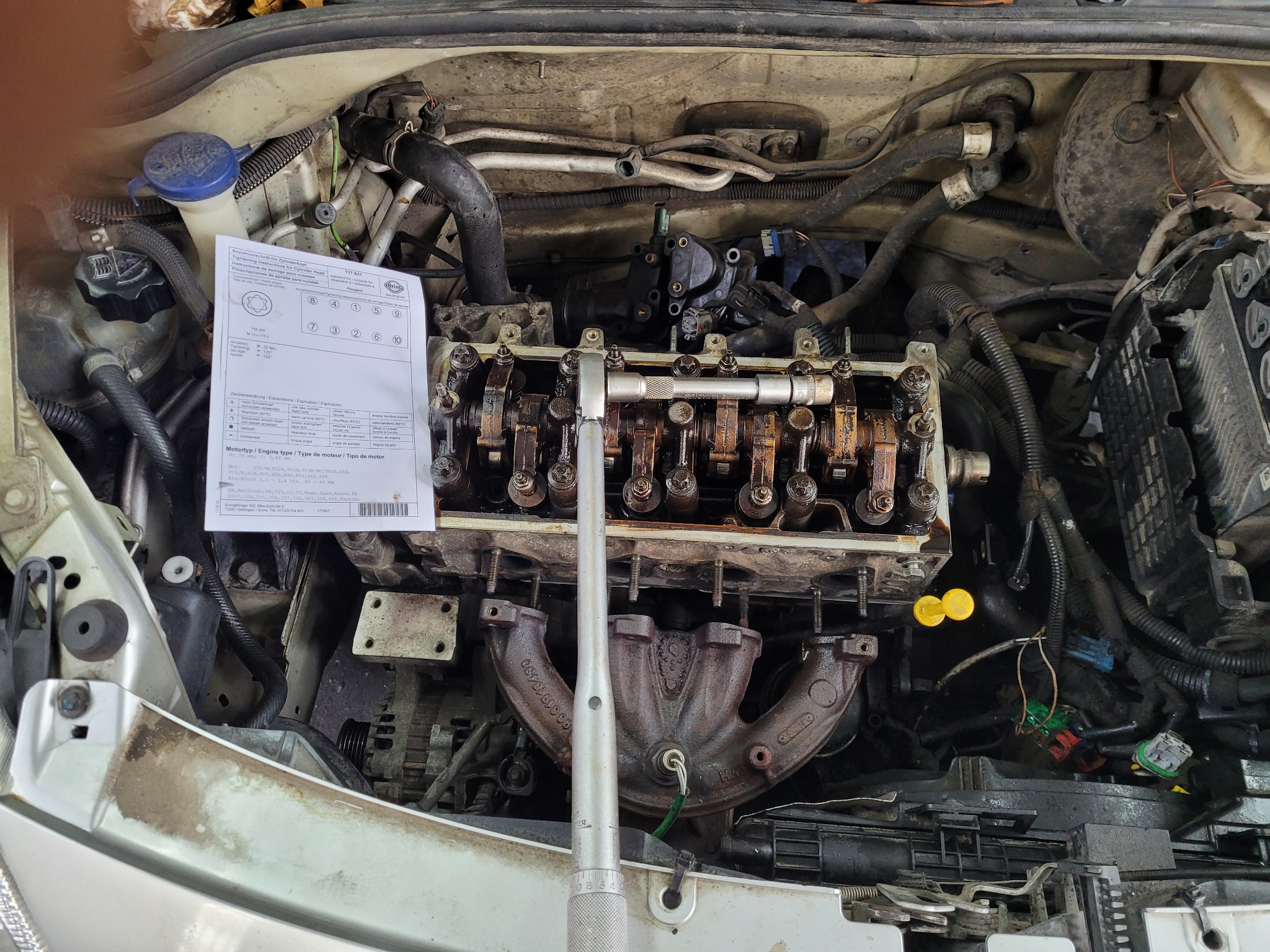 Silnik samochodowy z częściowo zdemontowaną głowicą cylindrów, widoczny klucz dynamometryczny, instrukcja dokręcania śrub głowicy oraz zabrudzenia eksploatacyjne.