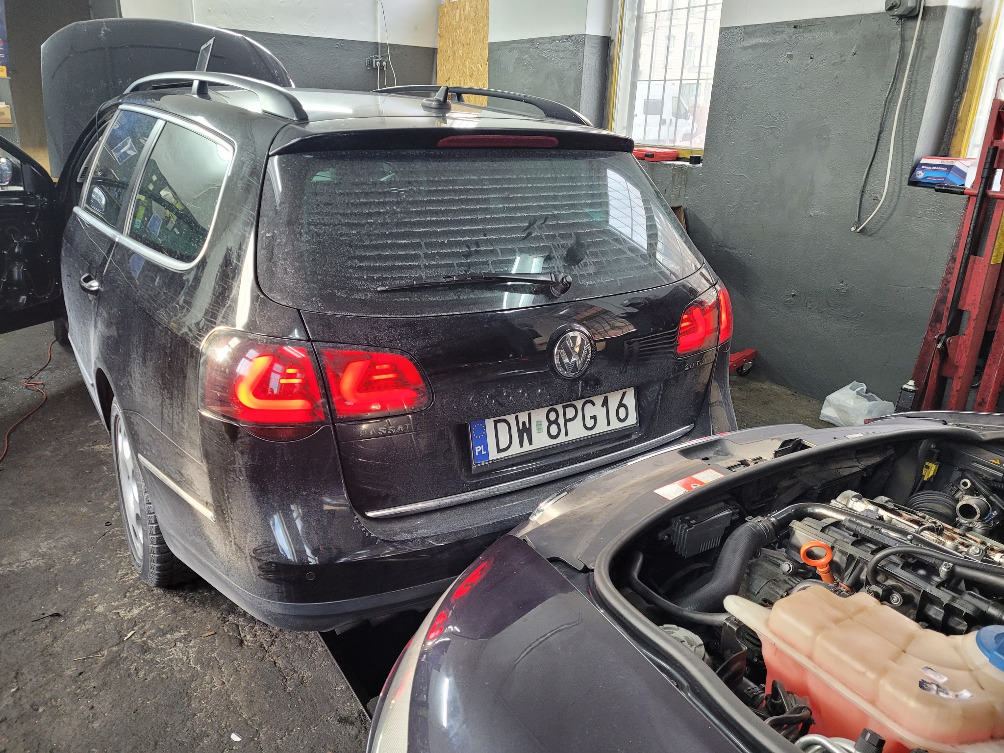 Czarny, zakurzony samochód kombi z otwartą klapą bagażnika, zaparkowany w warsztacie obok innego auta z otwartą maską, ukazującą silnik.