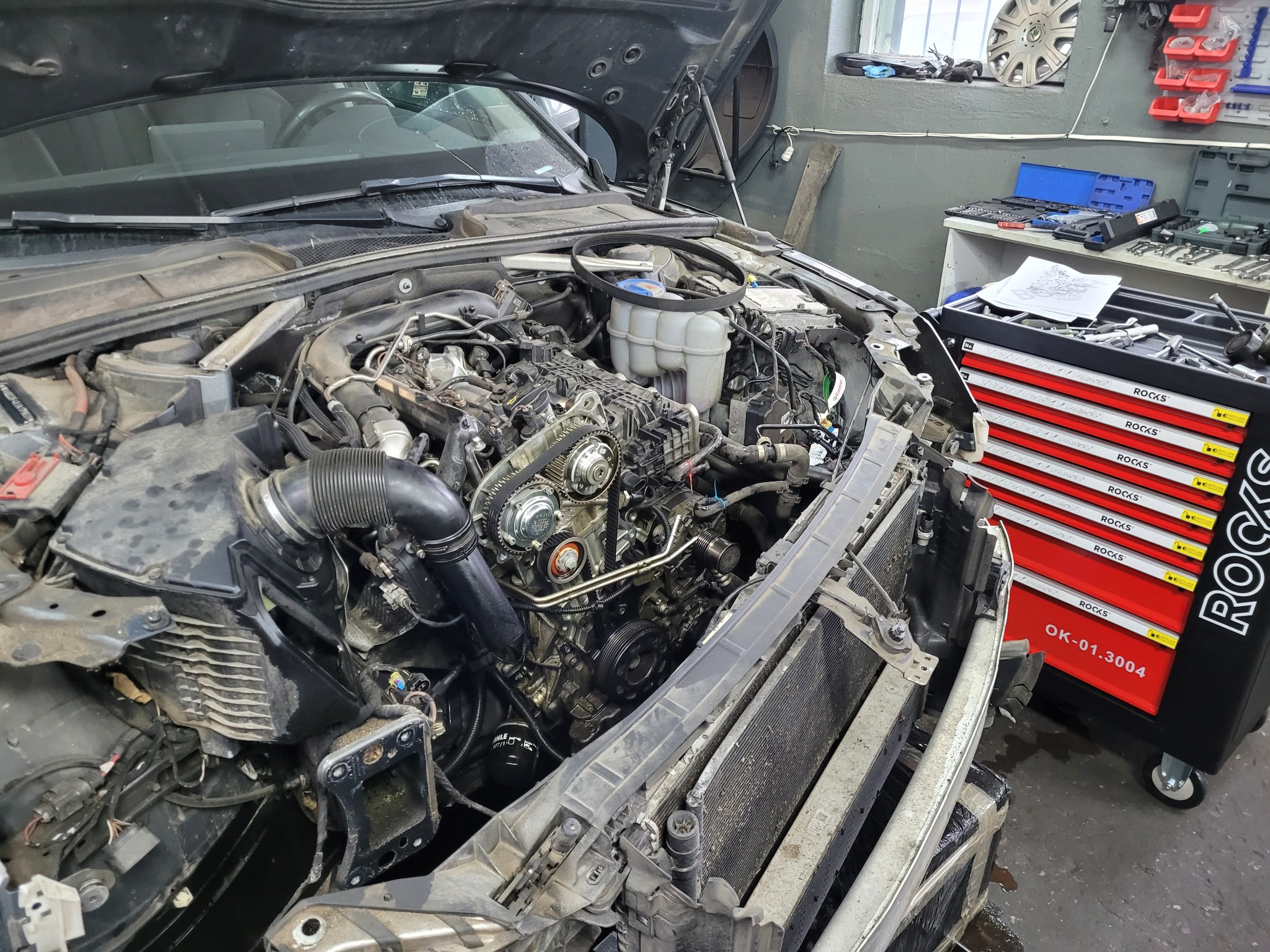 Widok z góry na otwartą maskę samochodu z widocznym silnikiem w trakcie naprawy, z elementami demontażu i skrzynką narzędziową 'ROCKS' w tle.