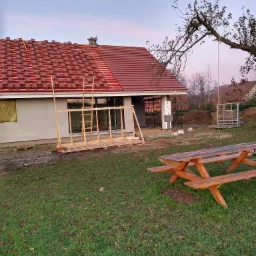 Dom w trakcie budowy z częściowo ułożoną czerwoną dachówką, widoczny fragment konstrukcji dachu, rusztowania i drewniany stół piknikowy na trawniku.