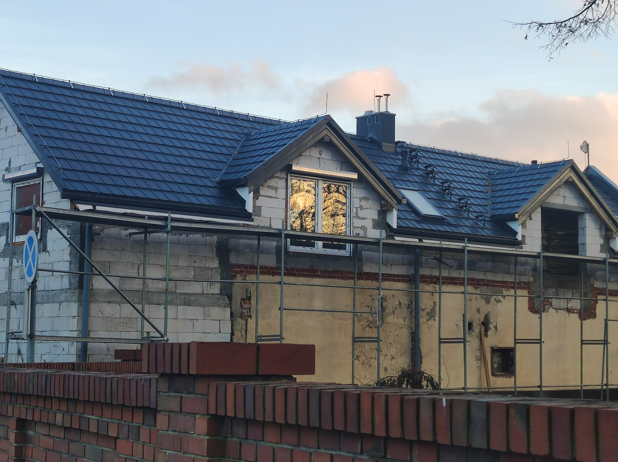 Dom w trakcie budowy z nowym, ciemnoszarym dachem z dachówki, widoczne rusztowania i ceglany mur w pierwszym planie.