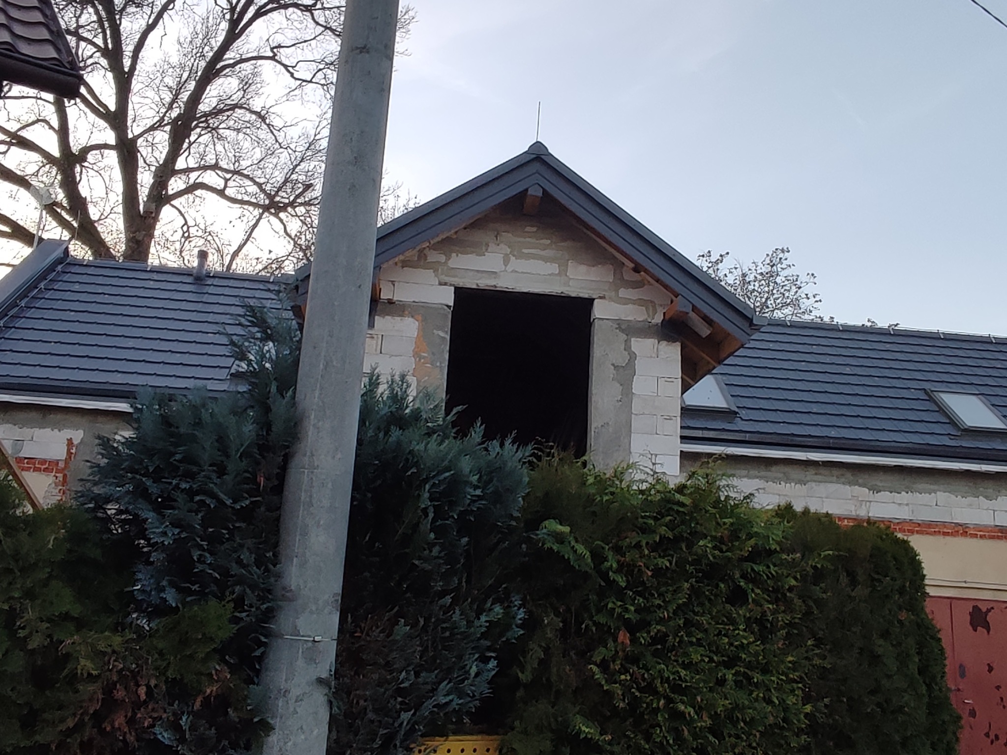 Surowy stan poddasza z nowym dachem z dachówki ceramicznej, widoczne bloczki betonowe i brakujące okno, na pierwszym planie gęste, zielone krzewy.