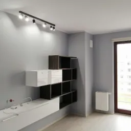 Szare wnętrze z nowoczesną zabudową meblową: białe i ciemnobrązowe szafki wiszące, listwa z reflektorami na suficie, grzejnik pod oknem balkonowym.