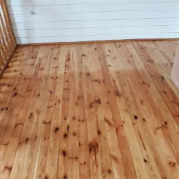 Drewniana podłoga z jasnych, lakierowanych paneli w domu w Lesznie, widok z perspektywy podłogi w kierunku ściany z boazerią i fragmentu kominka.