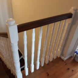 Widok z góry na drewniane schody z białymi balustradami i brązową poręczą; na podłodze stoi puszka z farbą i narzędzia malarskie.