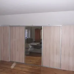 Wnętrze pokoju z dużą, jasną szafą wnękową z przesuwnymi drzwiami z lustrem, drewniana podłoga, widoczne gniazdko elektryczne na ścianie.