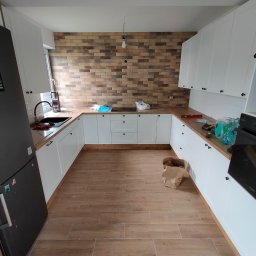 ŁUKASZ PANTKOWSKI - Nowoczesna kuchnia z białymi szafkami, drewnianym blatem i ceglaną ścianą w tle. Na blacie widoczne narzędzia i akcesoria kuchenne, a na podłodze kartonowe pudło.