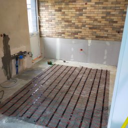 ŁUKASZ PANTKOWSKI - Montaż maty grzewczej pod podłogę w trakcie remontu, widoczne przygotowane ściany, częściowo ułożona cegła dekoracyjna i laser krzyżowy.