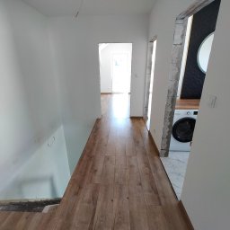 ŁUKASZ PANTKOWSKI - Korytarz w trakcie układania paneli podłogowych, widoczne niedokończone fragmenty przy schodach i w przejściach, białe ściany i pralka w tle.