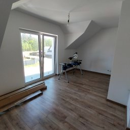 ŁUKASZ PANTKOWSKI - Pokój na poddaszu z ułożoną podłogą z paneli, widoczne paczki paneli pod oknem i piła do cięcia paneli na stojaku.