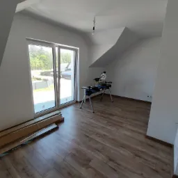 Pokój na poddaszu z ułożoną podłogą z paneli, widoczne paczki paneli pod oknem i piła do cięcia paneli na stojaku.