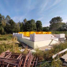ŁUKASZ PANTKOWSKI - Wznoszenie ścian fundamentowych z bloczków Ytong na ocieplonym fundamencie, widoczne żółte palety z bloczkami i stos metalowych rusztowań w trawie.
