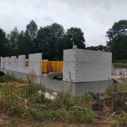 ŁUKASZ PANTKOWSKI - Wznoszenie ścian parteru domu z białych bloczków betonowych na wylanym fundamencie, widoczne żółte palety z materiałem budowlanym i otaczająca działkę zieleń.