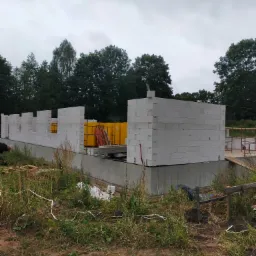 Wznoszenie ścian parteru domu z białych bloczk&oacute;w betonowych na wylanym fundamencie, widoczne ż&oacute;łte palety z materiałem budowlanym i otaczająca działkę zieleń.