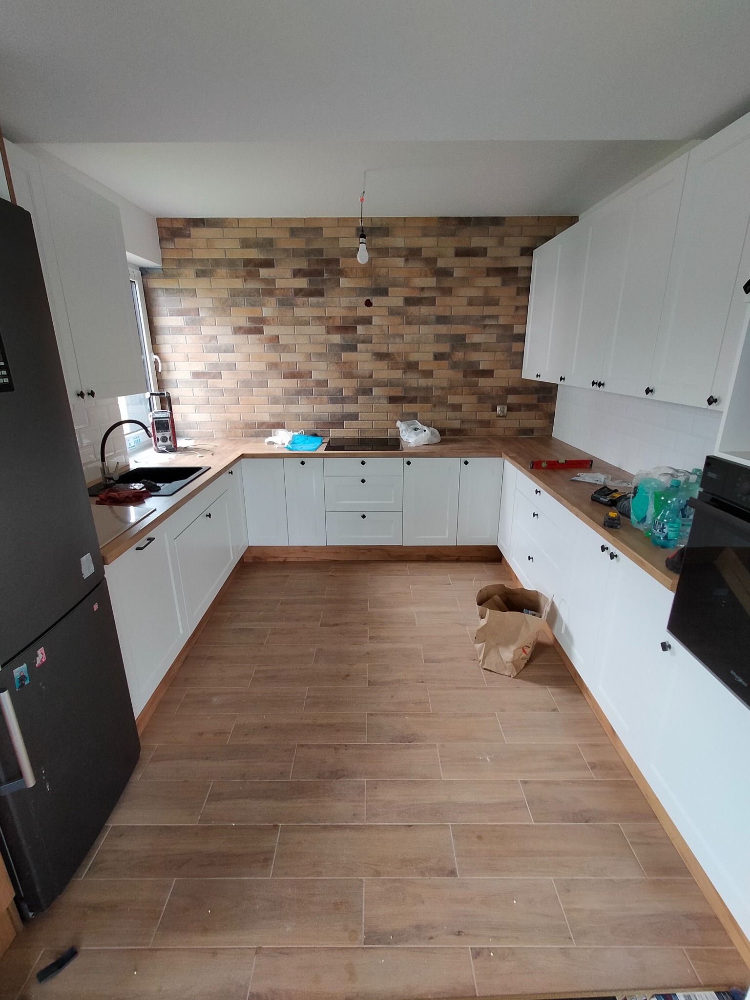 Nowoczesna kuchnia z białymi szafkami, drewnianym blatem i ceglaną ścianą w tle. Na blacie widoczne narzędzia i akcesoria kuchenne, a na podłodze kartonowe pudło.