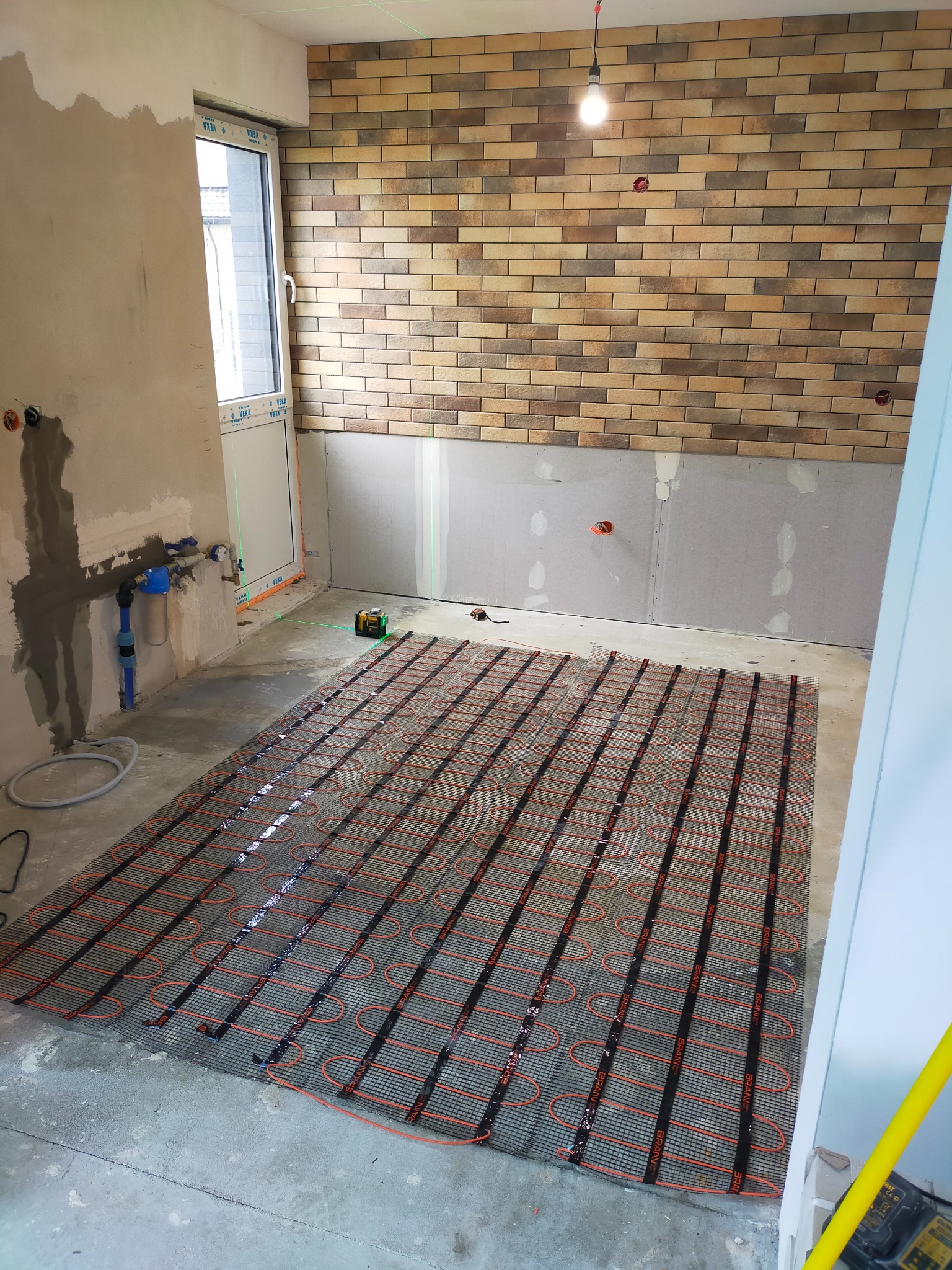 Montaż maty grzewczej pod podłogę w trakcie remontu, widoczne przygotowane ściany, częściowo ułożona cegła dekoracyjna i laser krzyżowy.