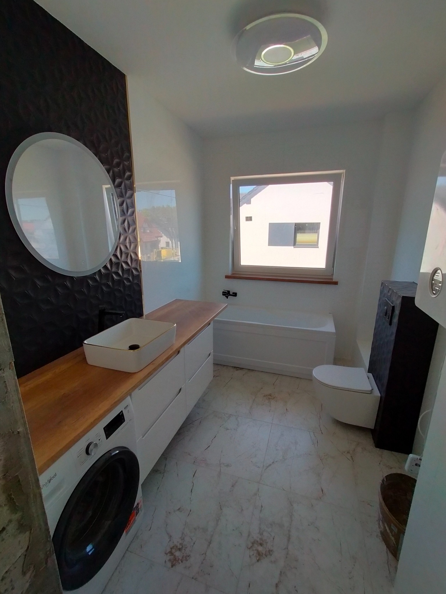 Nowoczesna łazienka z umywalką nablatową, drewnianym blatem, pralką Whirlpool, czarną ścianą z geometrycznym wzorem i okrągłym lustrem.