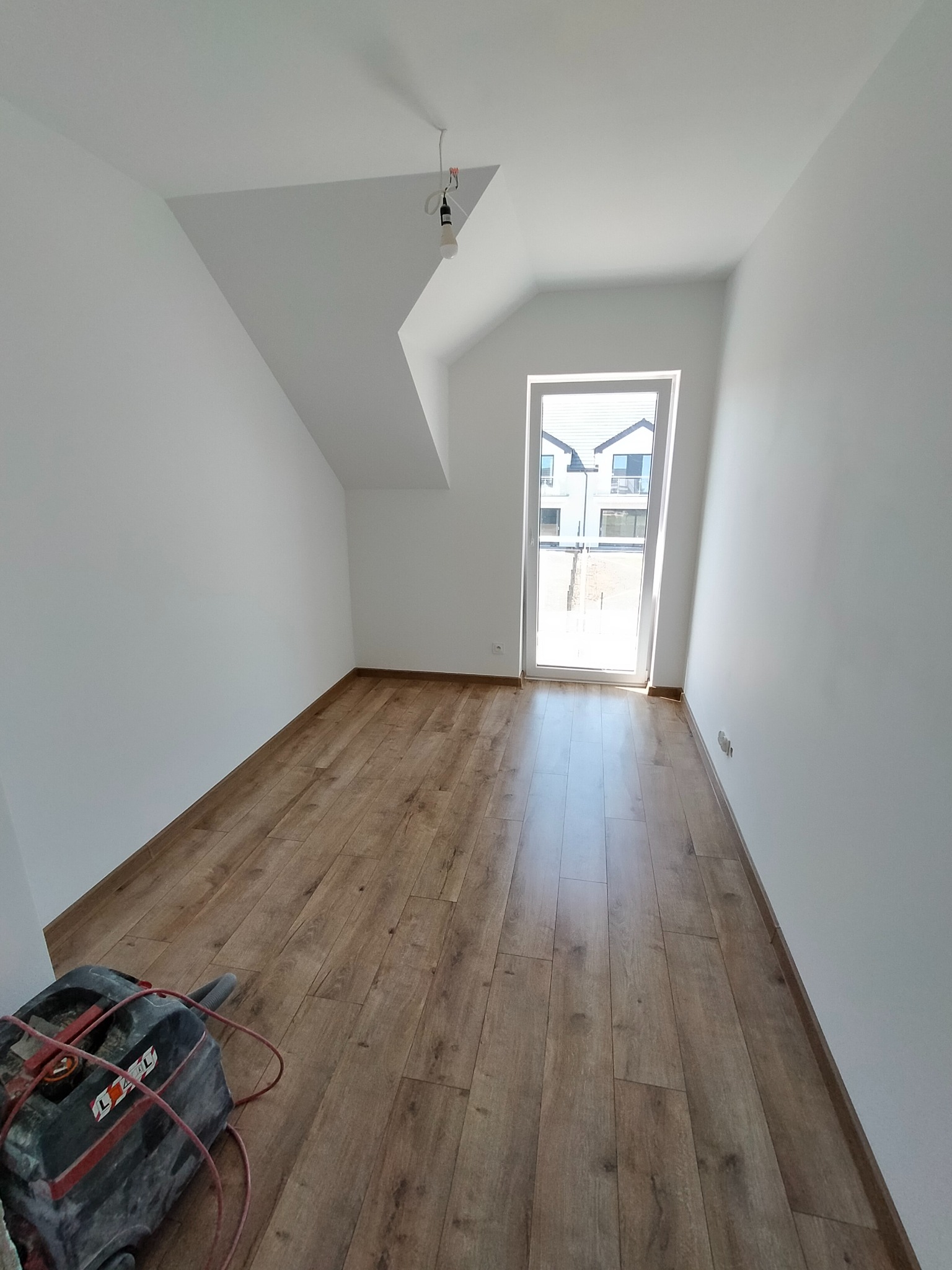 Pokój na poddaszu z nowo ułożonymi panelami podłogowymi o wzorze drewna, widoczne białe ściany, okno balkonowe i odkurzacz przemysłowy w rogu pomieszczenia.
