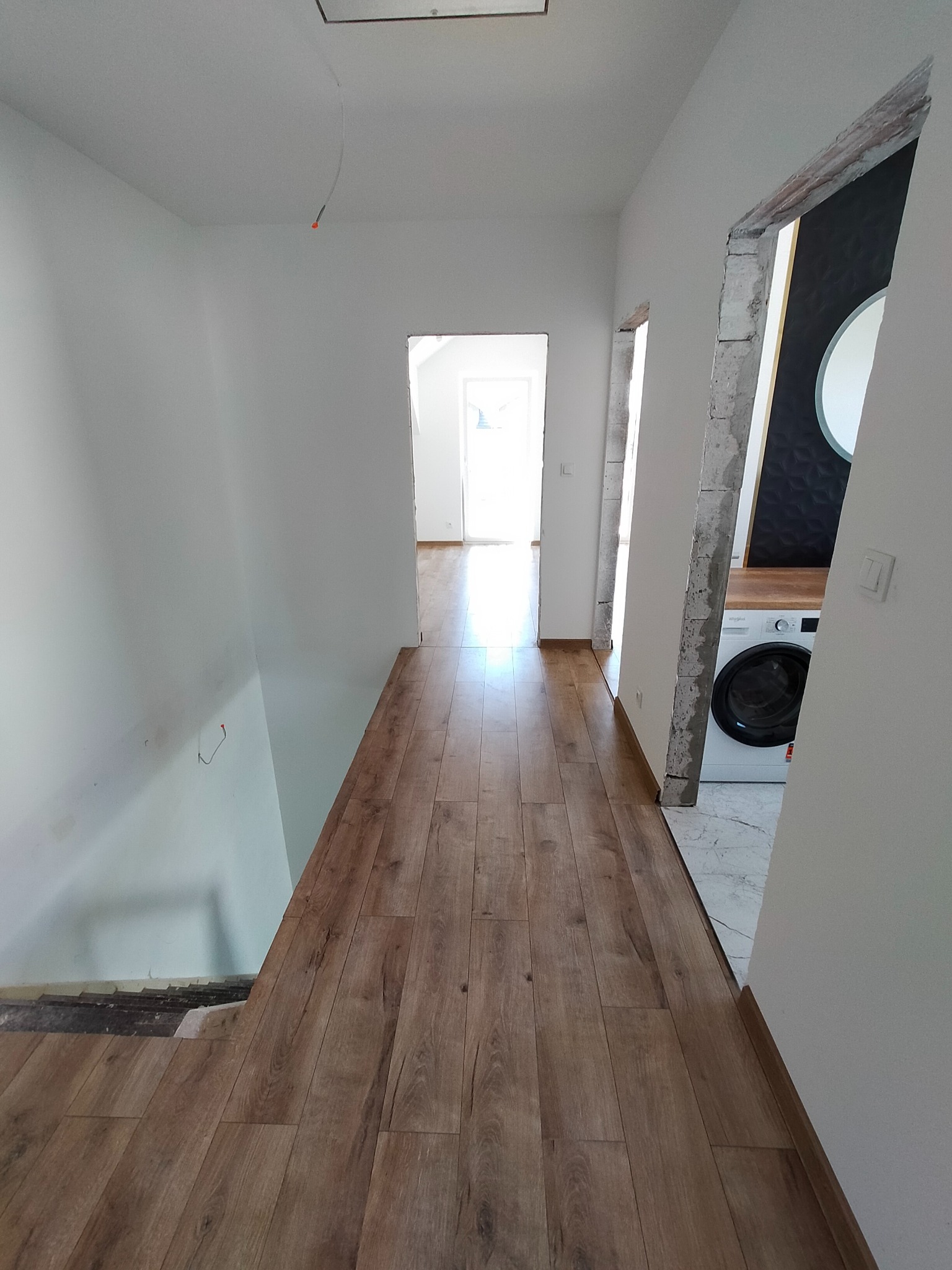 Korytarz w trakcie układania paneli podłogowych, widoczne niedokończone fragmenty przy schodach i w przejściach, białe ściany i pralka w tle.