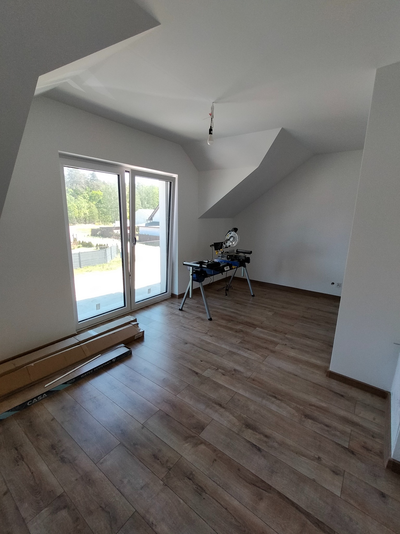 Pokój na poddaszu z ułożoną podłogą z paneli, widoczne paczki paneli pod oknem i piła do cięcia paneli na stojaku.