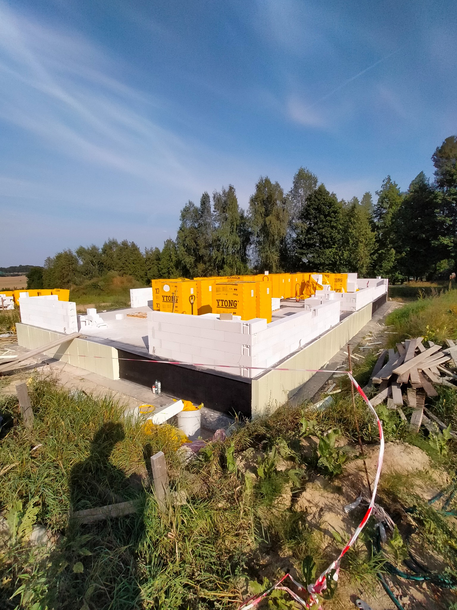 Budowa domu jednorodzinnego: fundamenty z izolacją i pierwsze warstwy ścian z bloczków Ytong, widoczne palety z materiałem, teren budowy zabezpieczony taśmą.