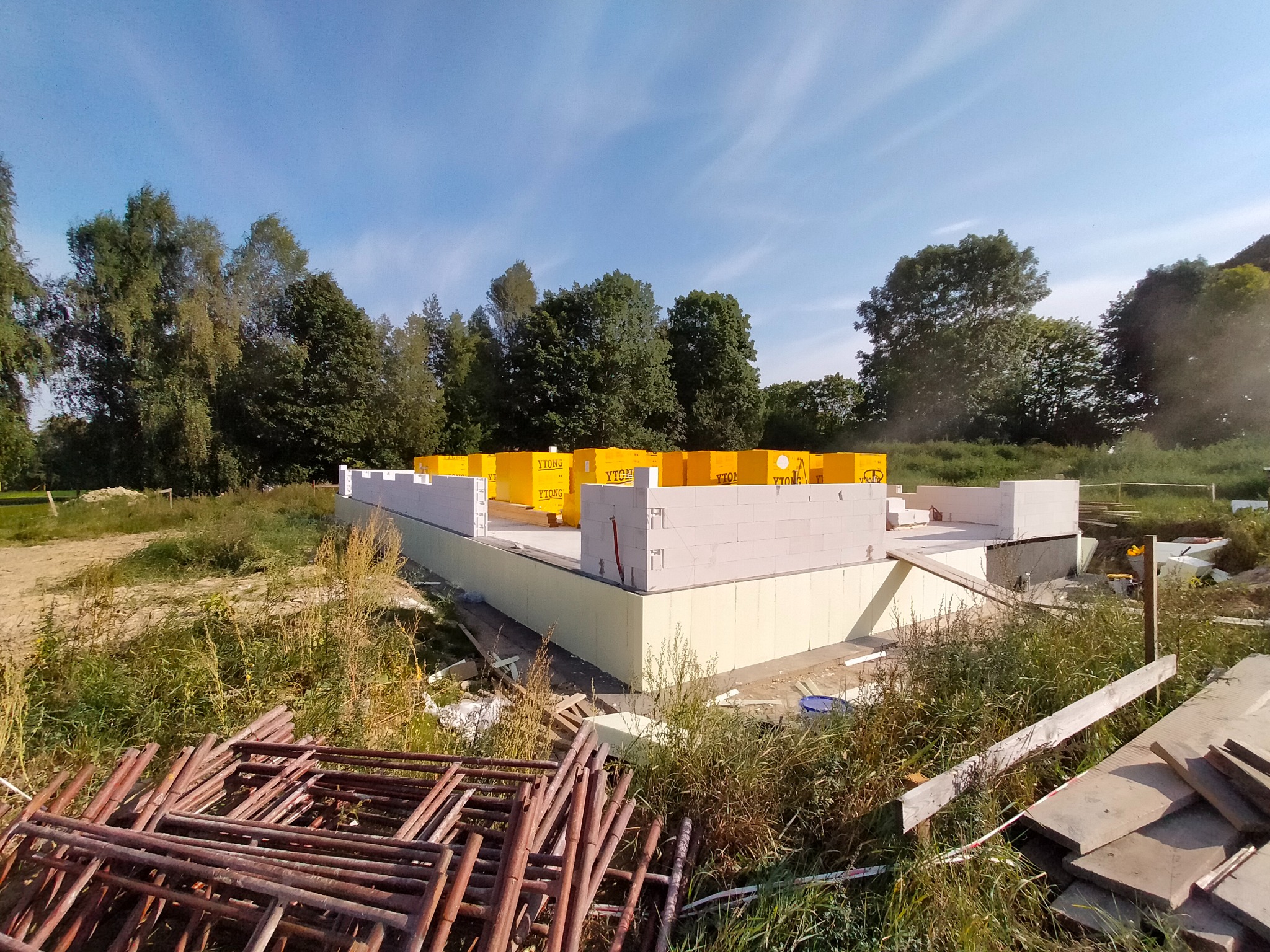 Wznoszenie ścian fundamentowych z bloczków Ytong na ocieplonym fundamencie, widoczne żółte palety z bloczkami i stos metalowych rusztowań w trawie.