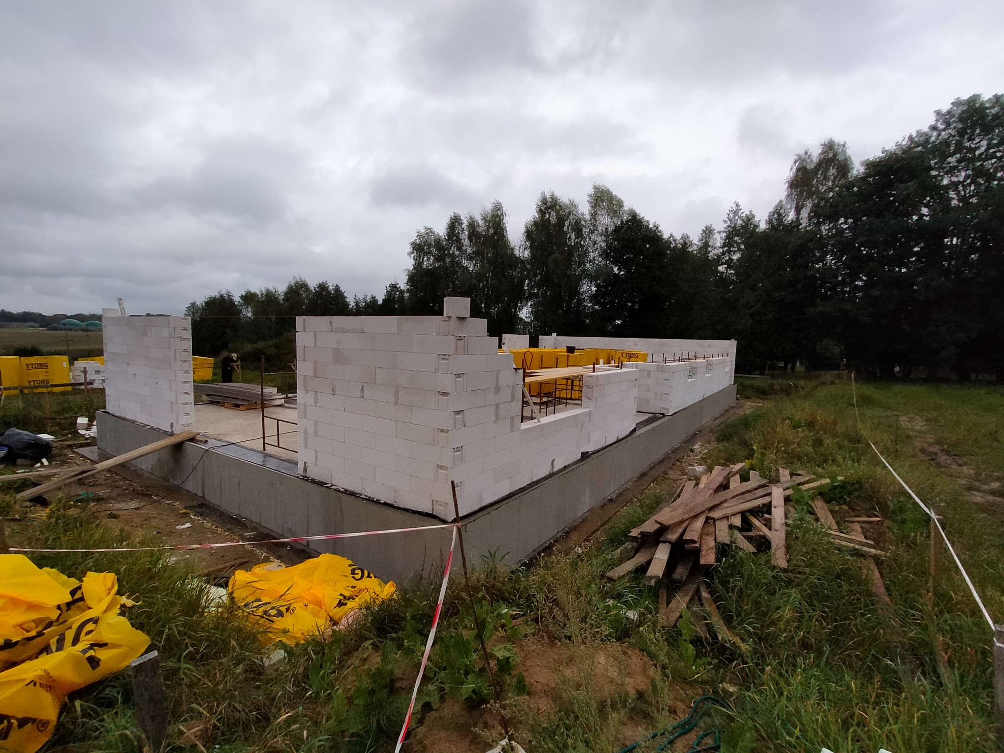 Wznoszone ściany domu z białych bloczków Ytong na betonowym fundamencie, widoczne żółte opakowania bloczków i rozrzucone deski na zewnątrz, pochmurne niebo w tle.