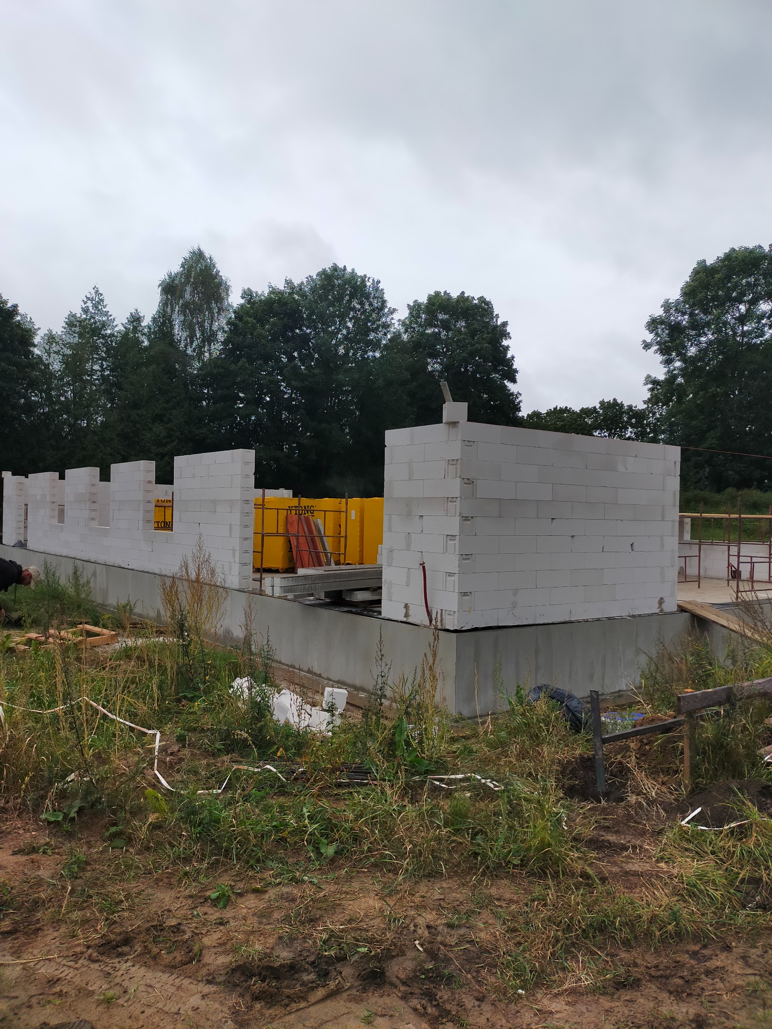 Wznoszenie ścian parteru domu z białych bloczków betonowych na wylanym fundamencie, widoczne żółte palety z materiałem budowlanym i otaczająca działkę zieleń.