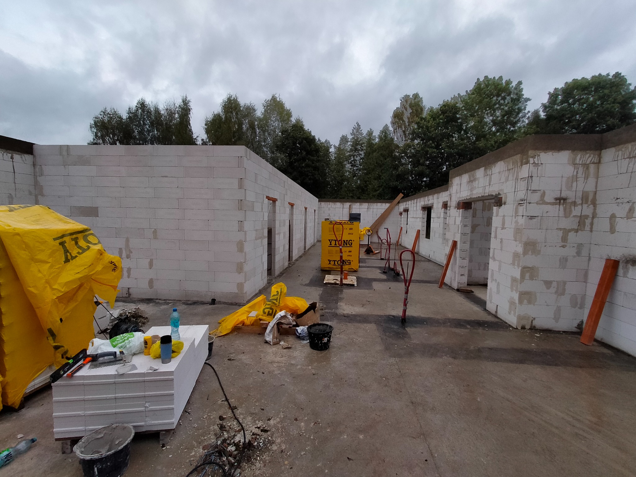 Wnętrze budynku w trakcie budowy z widocznymi ścianami z białych bloczków, paletą bloczków Ytong, narzędziami murarskimi, folią ochronną i elementami konstrukcyjnymi w szary, pochmurny dzień.