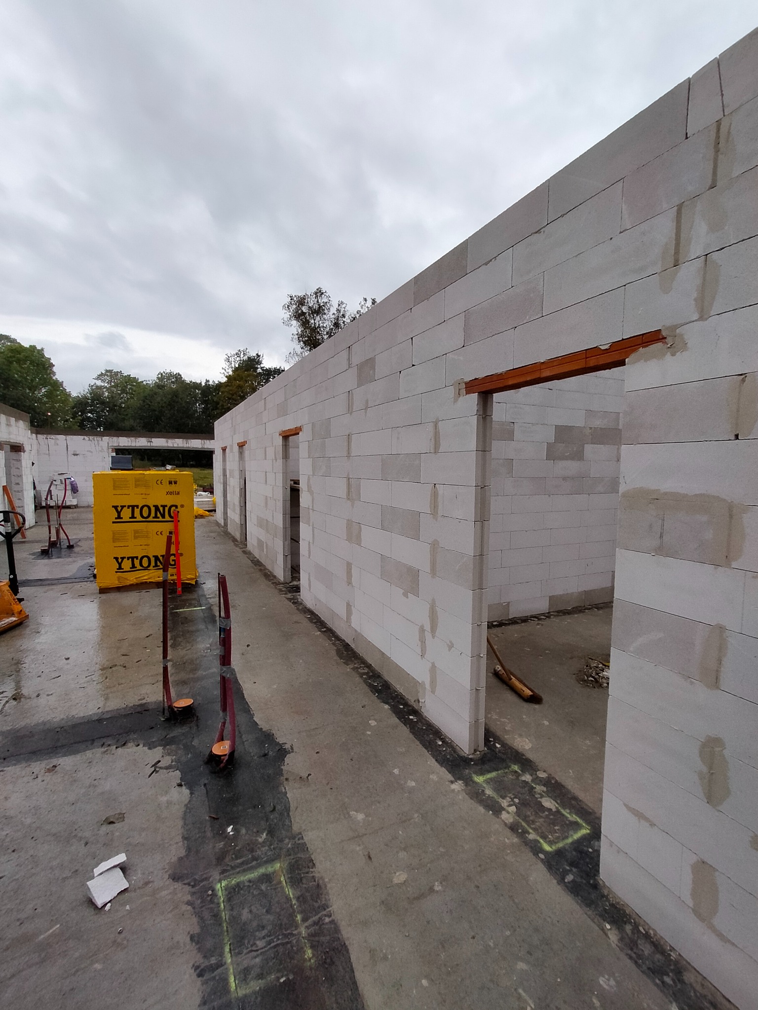 Wznoszenie ścian z bloczków Ytong: surowy stan budynku z widocznymi otworami drzwiowymi, paleta bloczków i narzędzia budowlane na mokrej posadzce.