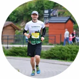 Uśmiechnięty mężczyzna w trakcie biegu ulicznego, ubrany w koszulkę sportową z numerem startowym, czarne spodenki i niebieskie buty. W tle widoczna zielona roślinność, drewniana chatka i fragment...