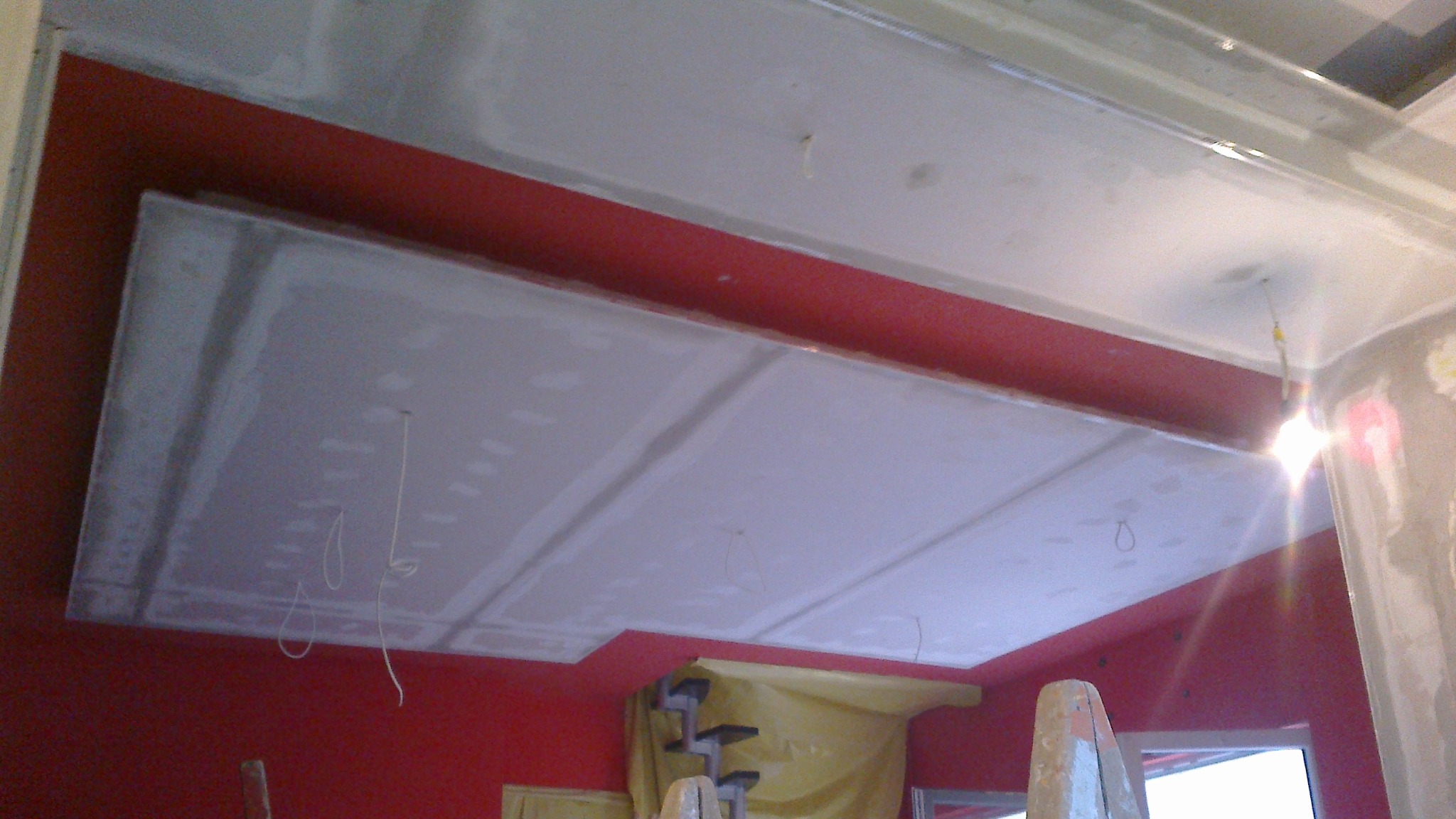 Czerwona ściana z zamontowanym sufitem podwieszanym z płyt kartonowo-gipsowych, widoczne szpachlowane łączenia płyt, w tle drabina malarska przykryta folią ochronną.