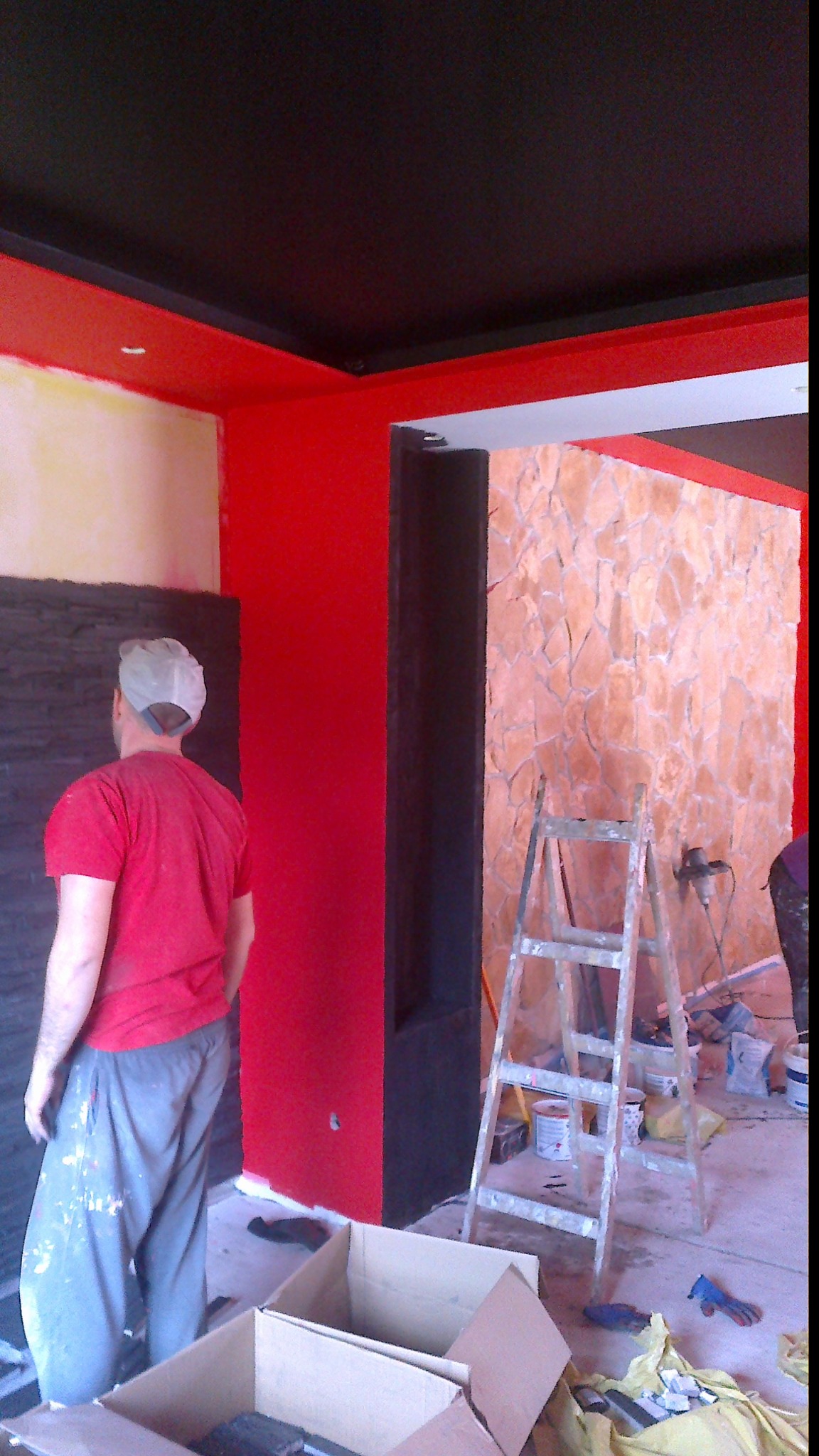 Mężczyzna w czerwonej koszulce stoi w pomieszczeniu z pomalowanymi na czerwono ścianami, czarnym sufitem i dekoracyjną ścianą z kamienia, widoczna drabina, kartony i narzędzia malarskie.
