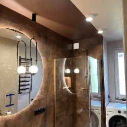 Nowoczesna łazienka z kabiną prysznicową typu walk-in, okrągłym lustrem z podświetleniem LED i wiszącymi lampami. Brązowe płytki imitujące rdzę. Pralka obok kabiny.