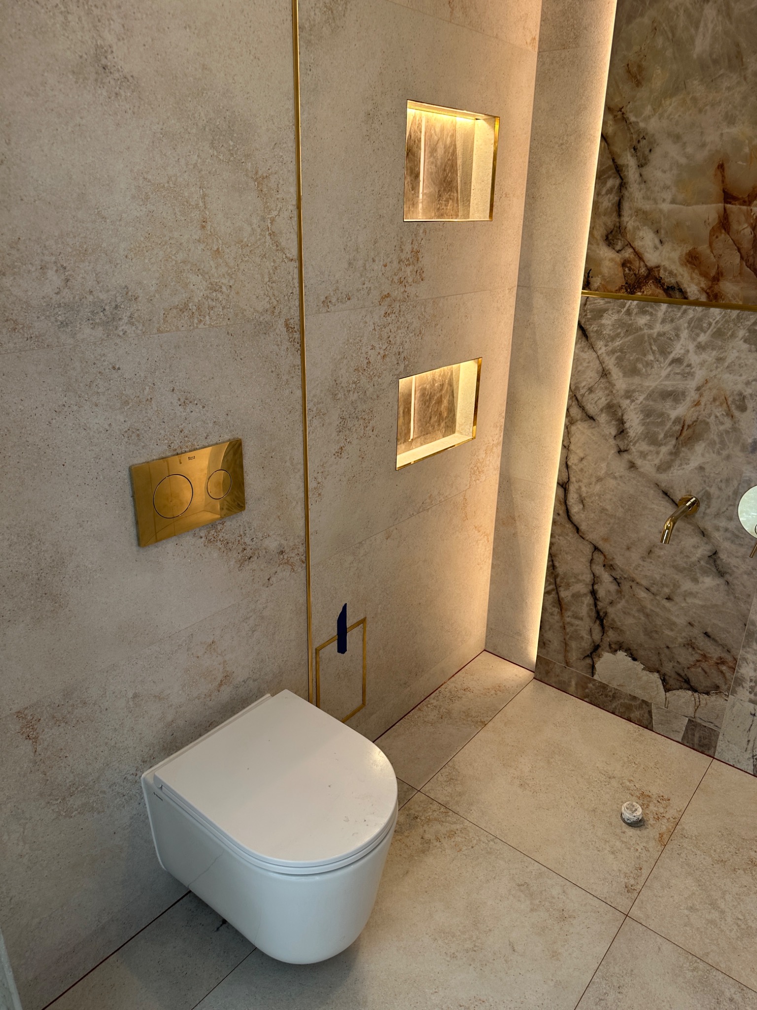Elegancka łazienka po remoncie: podwieszana toaleta, beżowe płytki z złotymi akcentami, oświetlone nisze w ścianie i marmurowa ściana z prysznicem.