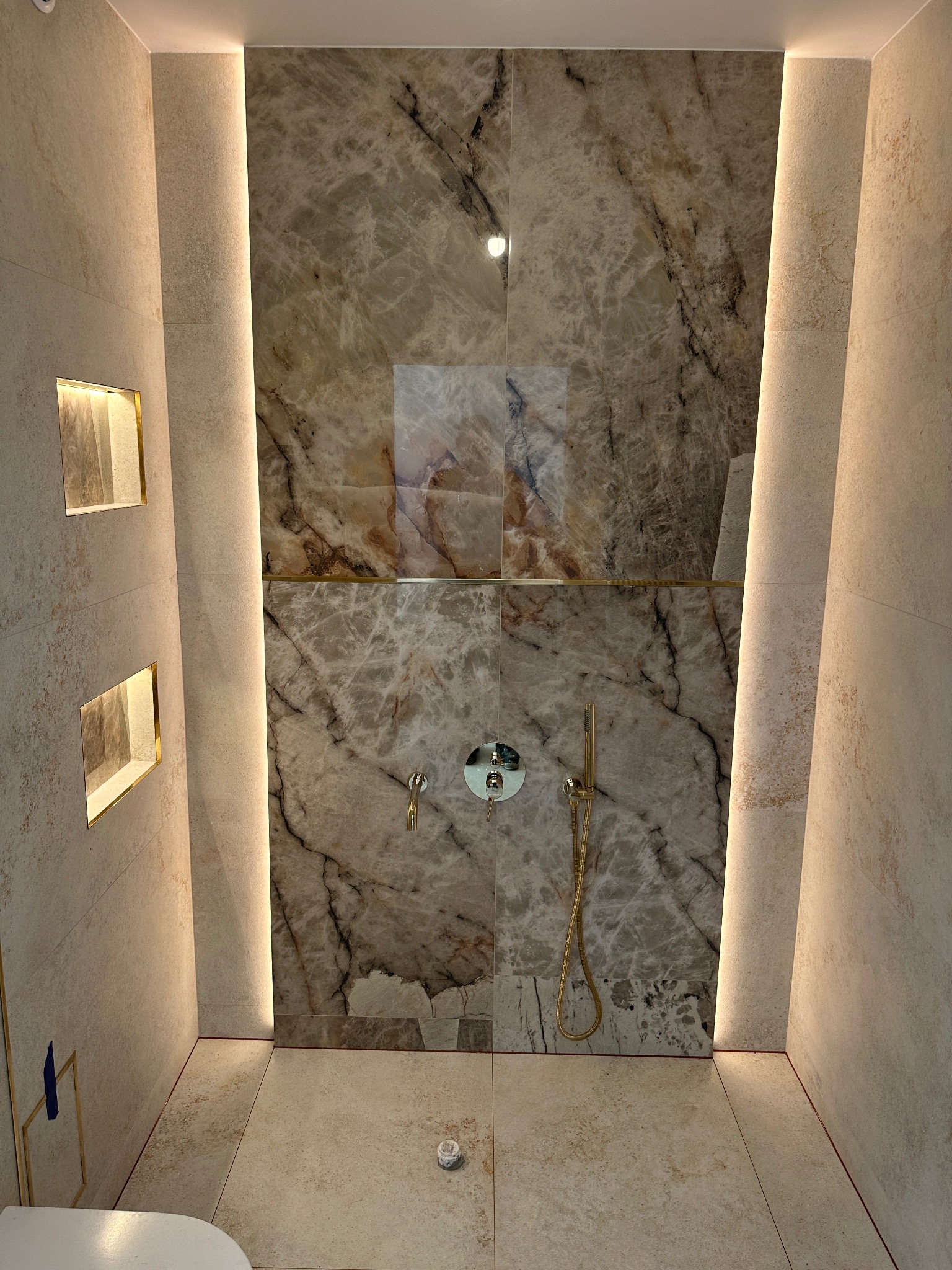Elegancka łazienka z marmurową ścianą prysznicową i złotą armaturą. Podświetlane wnęki i listwy LED tworzą ciepłą atmosferę. Widoczny fragment toalety.