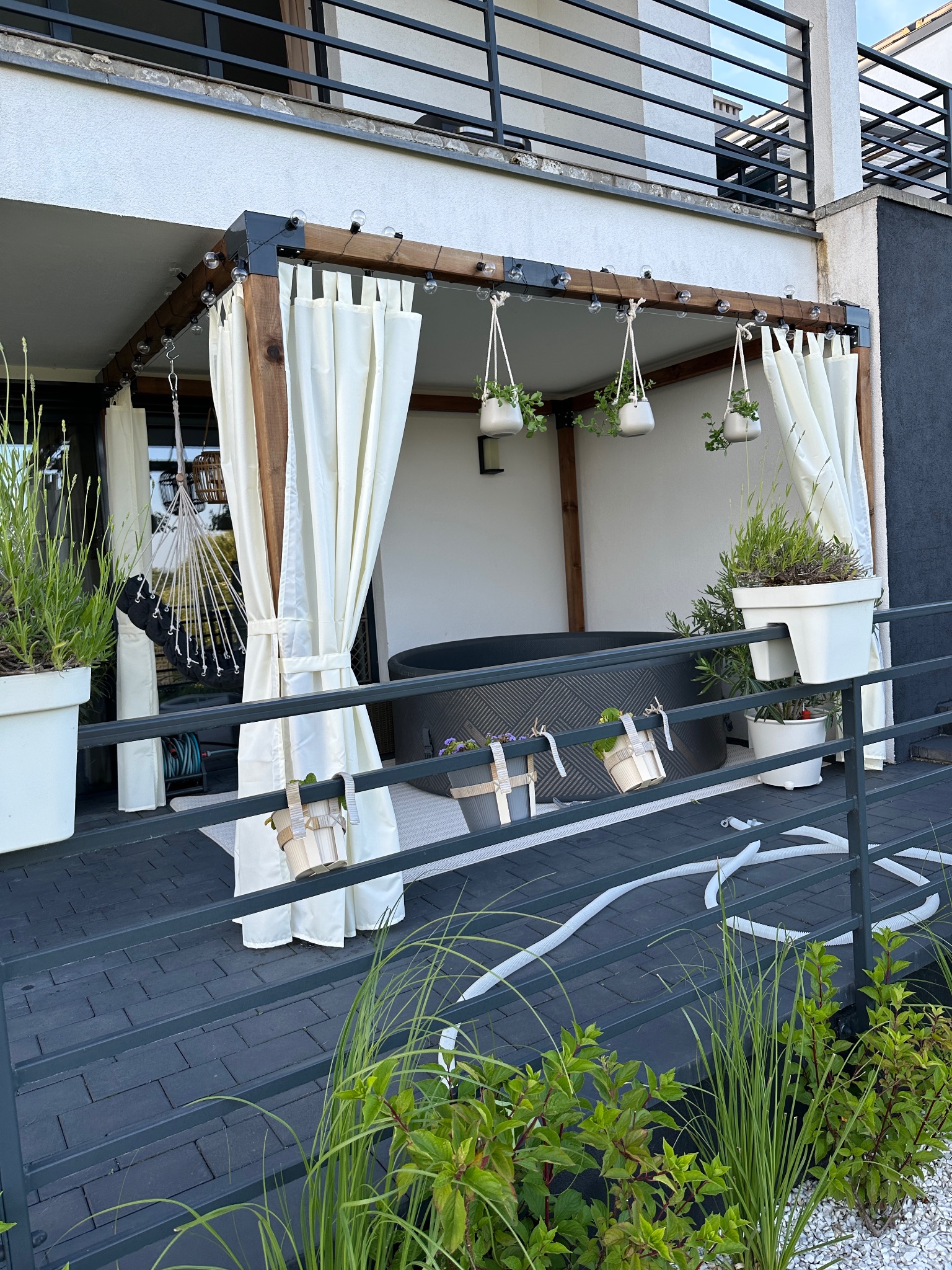 Zadaszony taras z jacuzzi, ozdobiony białymi zasłonami, wiszącymi doniczkami i roślinami w donicach na balustradzie. W tle widoczny budynek.