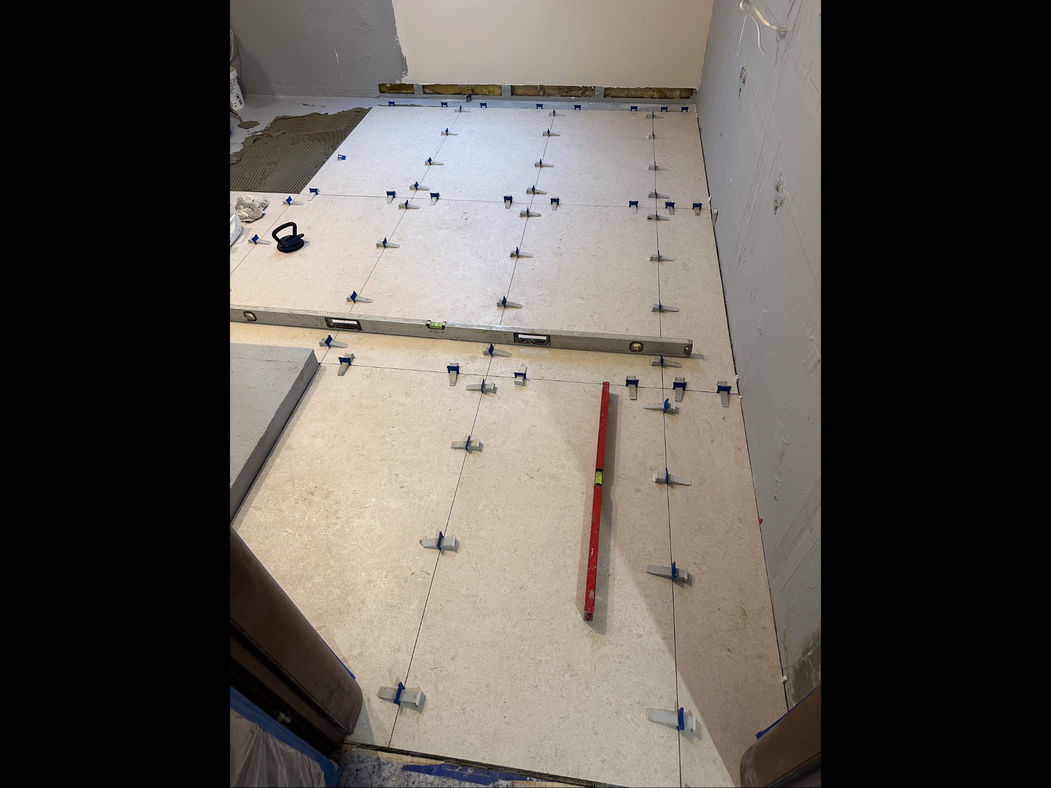 Układanie jasnych płytek podłogowych z użyciem poziomicy i klipsów. Częściowo położone płytki z klejem w tle. Remont w trakcie.