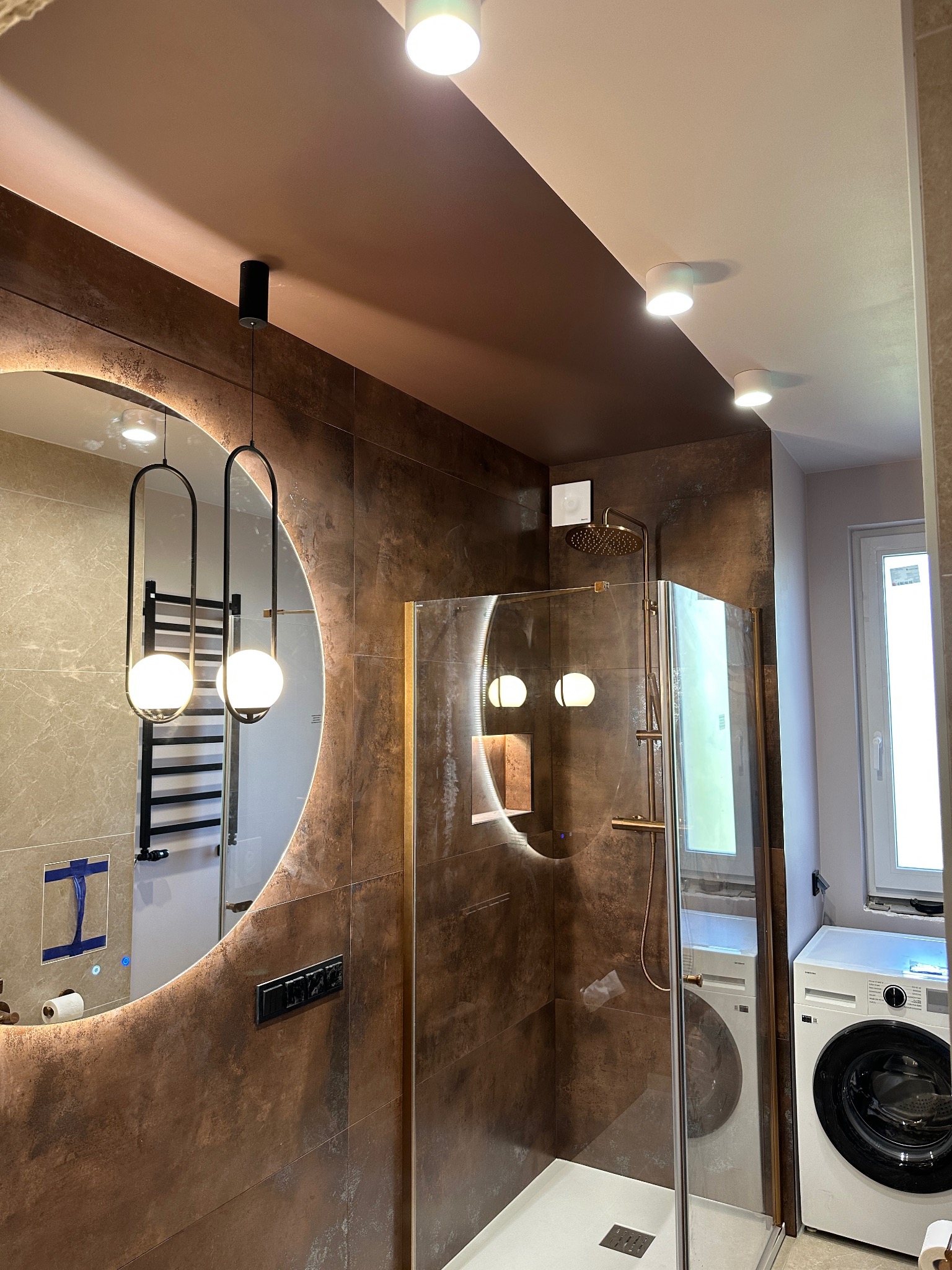 Nowoczesna łazienka z kabiną prysznicową typu walk-in, okrągłym lustrem z podświetleniem LED i wiszącymi lampami. Brązowe płytki imitujące rdzę. Pralka obok kabiny.
