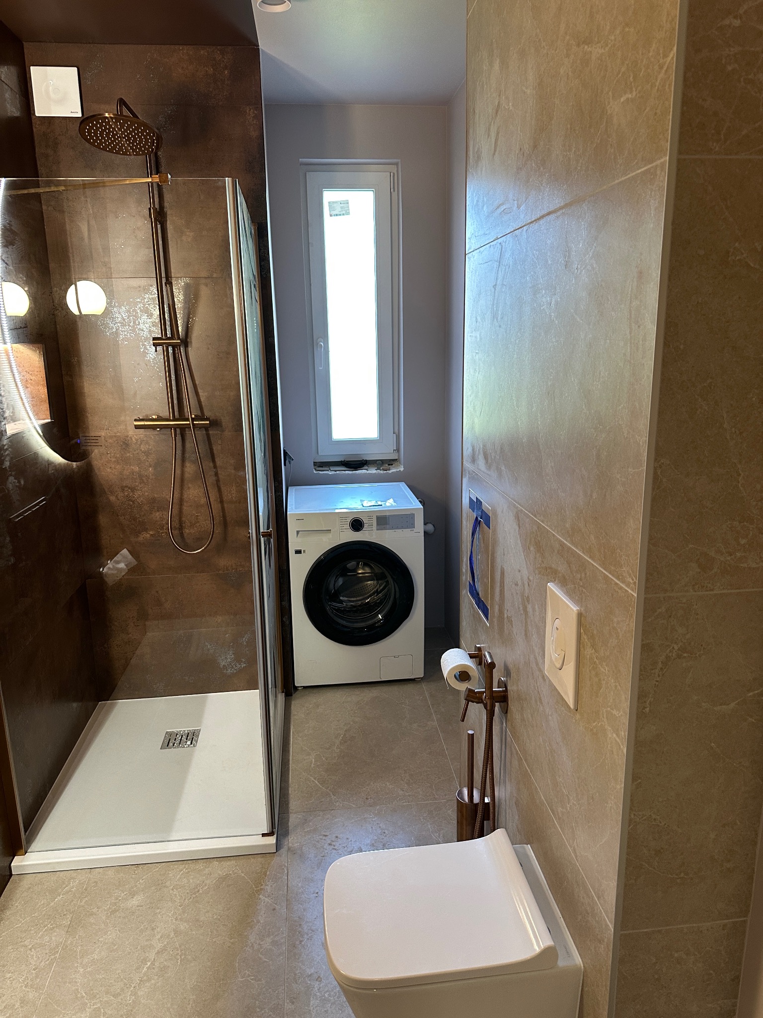 Nowoczesna łazienka z kabiną prysznicową typu walk-in, toaletą wiszącą i pralką pod oknem. Użyto płytek w odcieniach brązu i beżu. Armatura w kolorze miedzi.