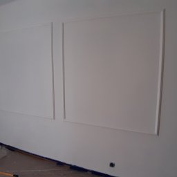 KBM Krzysztof Błażewicz - Świeżo pomalowana ściana z dekoracyjnymi ramkami, zabezpieczona folią podłoga, widoczny wałek malarski.