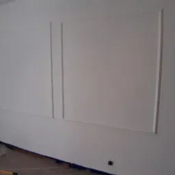 Świeżo pomalowana ściana z dekoracyjnymi ramkami, zabezpieczona folią podłoga, widoczny wałek malarski.