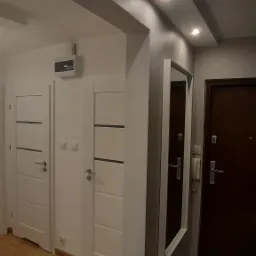 Korytarz po remoncie z dwiema białymi drzwiami z poziomymi frezowaniami, lustrem w białej ramie, parkietem i fragmentem marmurowej podłogi.