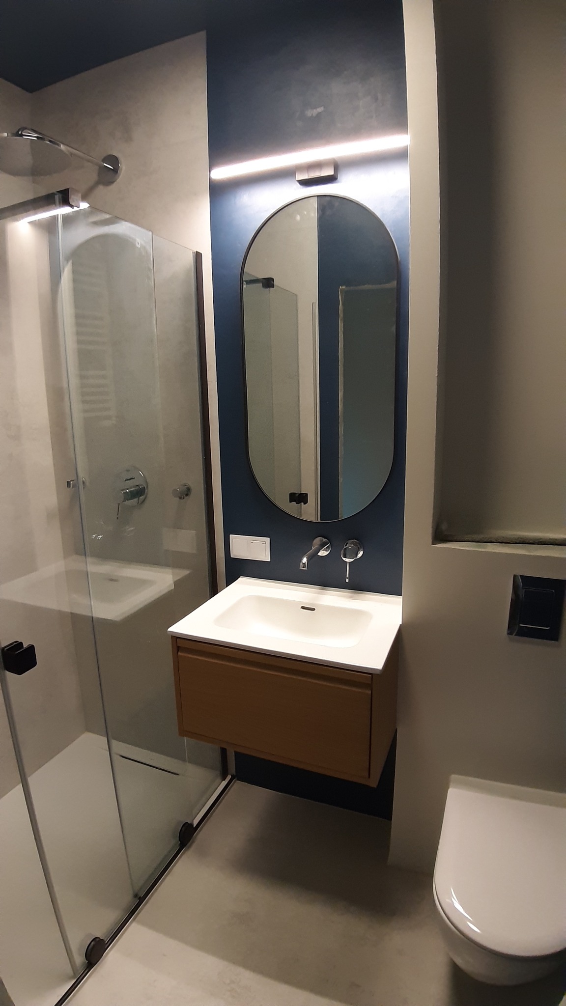 Nowoczesna łazienka z kabiną prysznicową ze szklanymi drzwiami, umywalką z szafką w kolorze drewna i owalnym lustrem podświetlonym listwą LED na ścianie w odcieniu granatu.