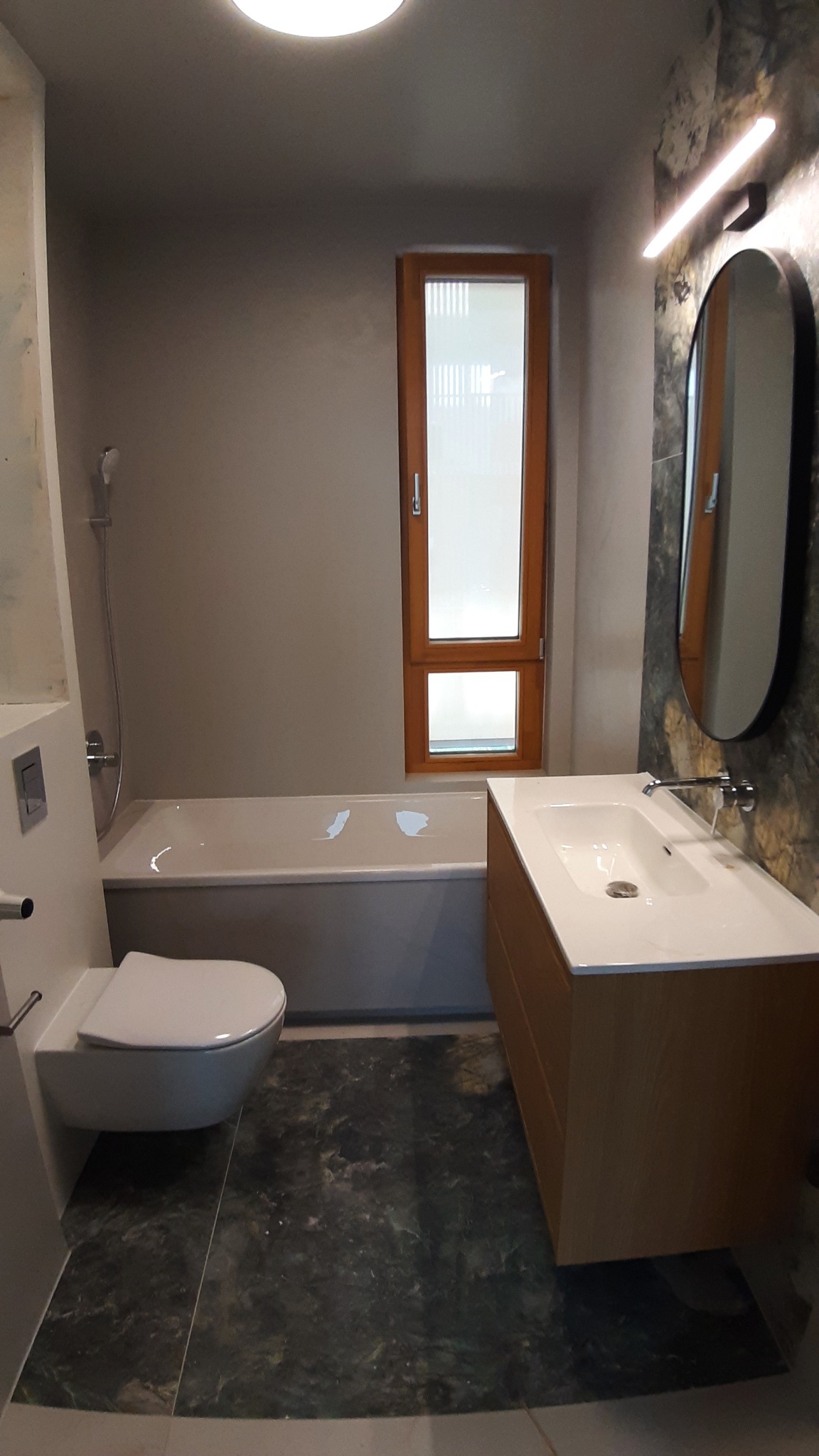 Nowoczesna łazienka z wanną, podwieszaną toaletą i umywalką na szafce z drewnianym fornirem, wykończona płytkami imitującymi marmur.