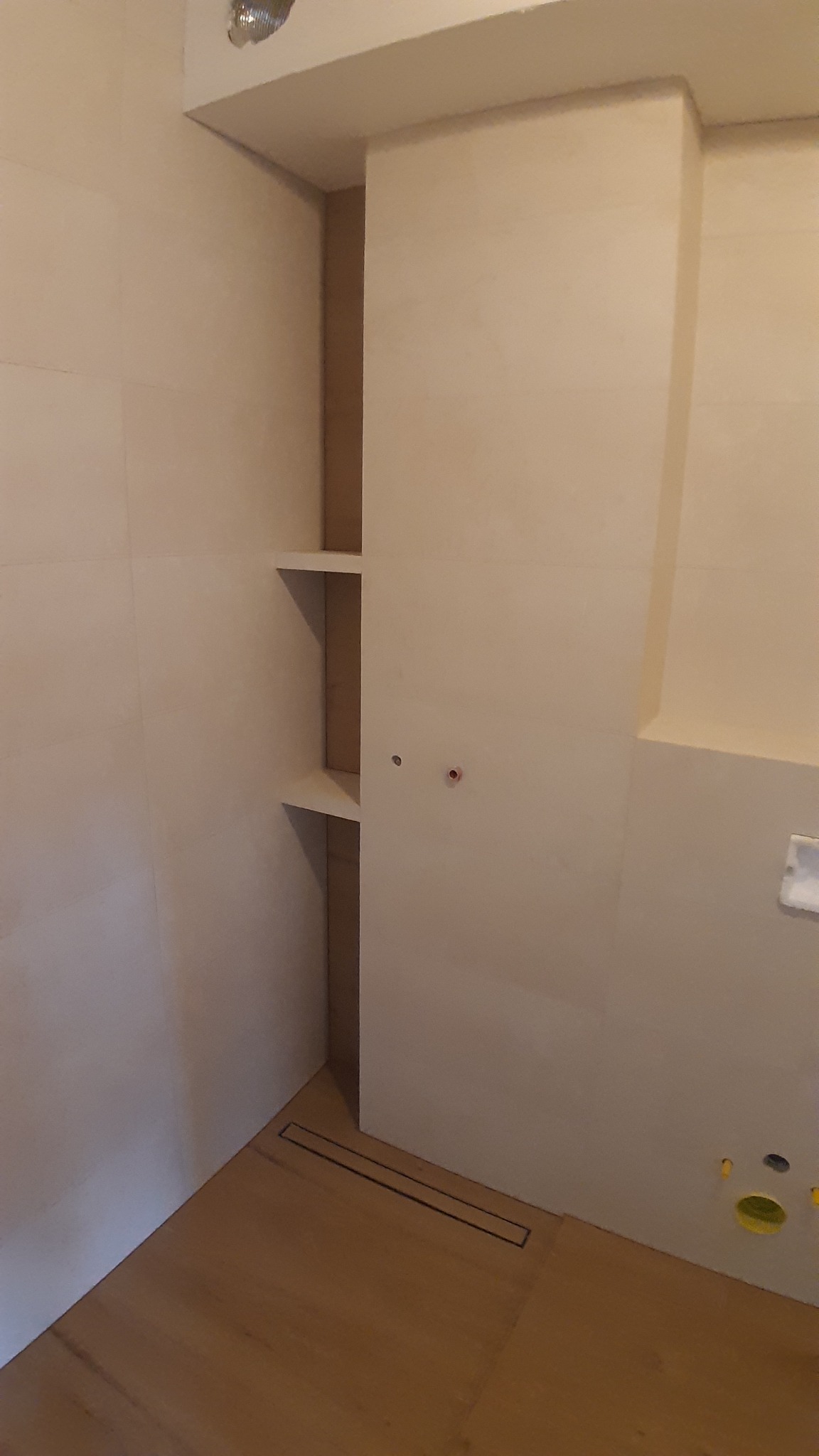 Beżowa łazienka w trakcie wykańczania, widoczna wnęka z półkami, odpływ liniowy w podłodze, przygotowane punkty instalacyjne na ścianie.